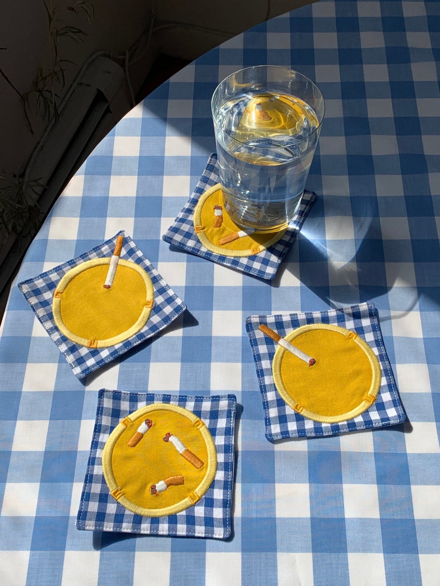 Set of 4 coasters photographed on top of a gingham blue tablecloth and a glass of water on one of them