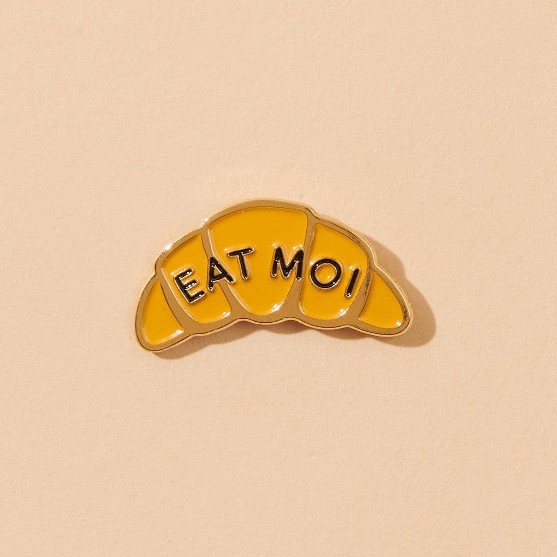 Enamel pin in the shape of a croissant that says "EAT MOI"