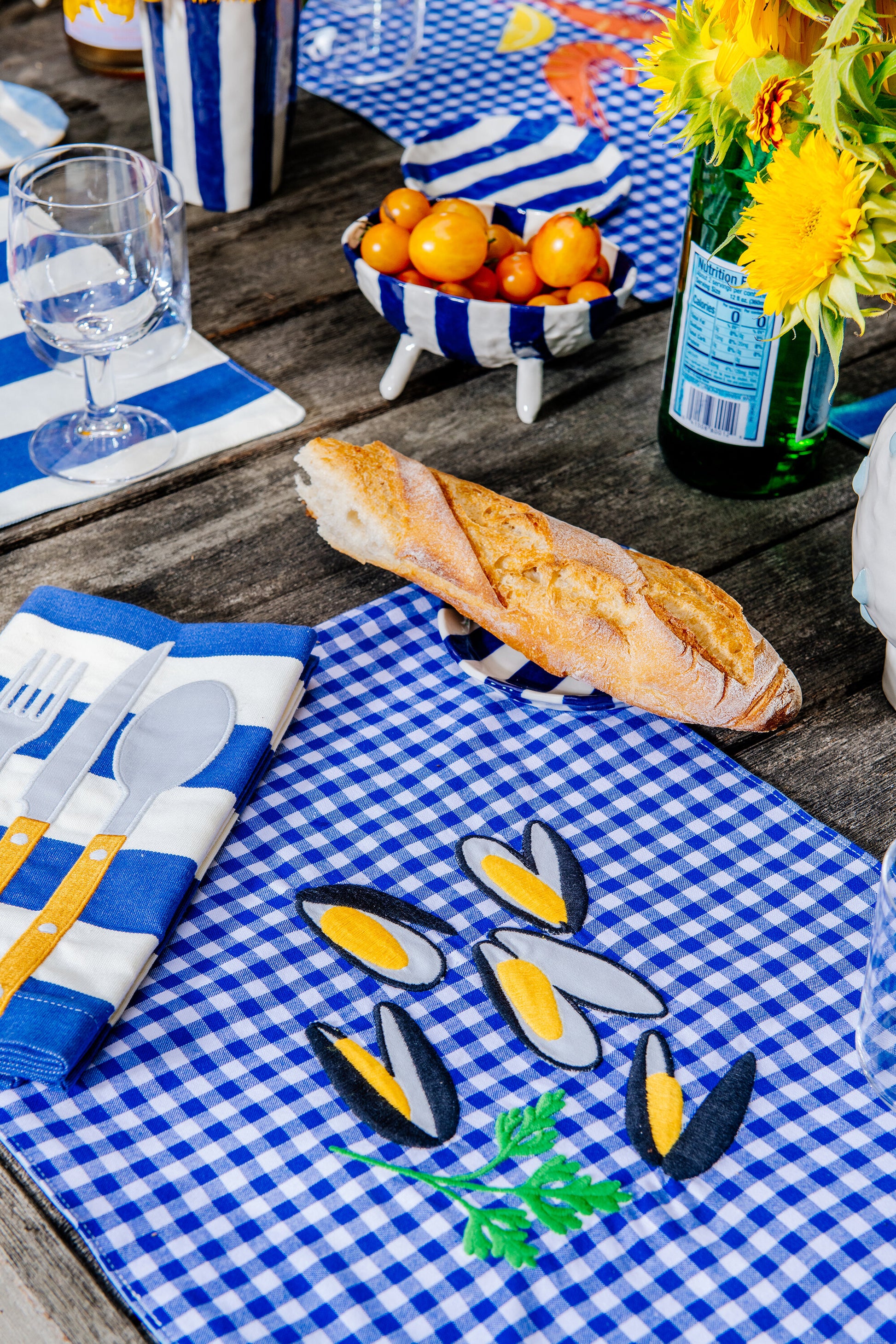 Table setting featuring a blue gingham placemat with five mussels and a parsley sprig embroidered in the center 