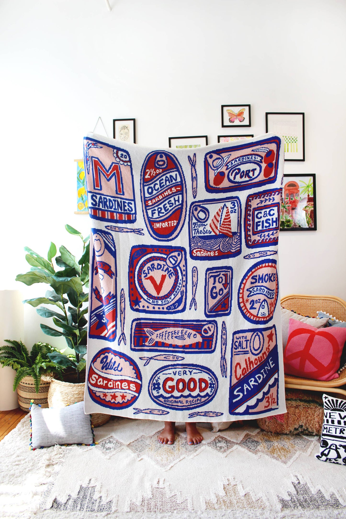 Sardines Knit Blanket in Mediterranean -- Various shaped sardine tin cans in soft pink, red, blue on an off white background colorway, held up by a person in their living room.