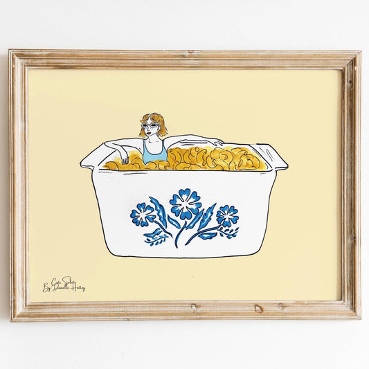 Wall print featuring an illustration of a girl sitting in a vintage CorningWare dish of Mac and Cheese with a pale yellow background. Shown in light colored wood frame.