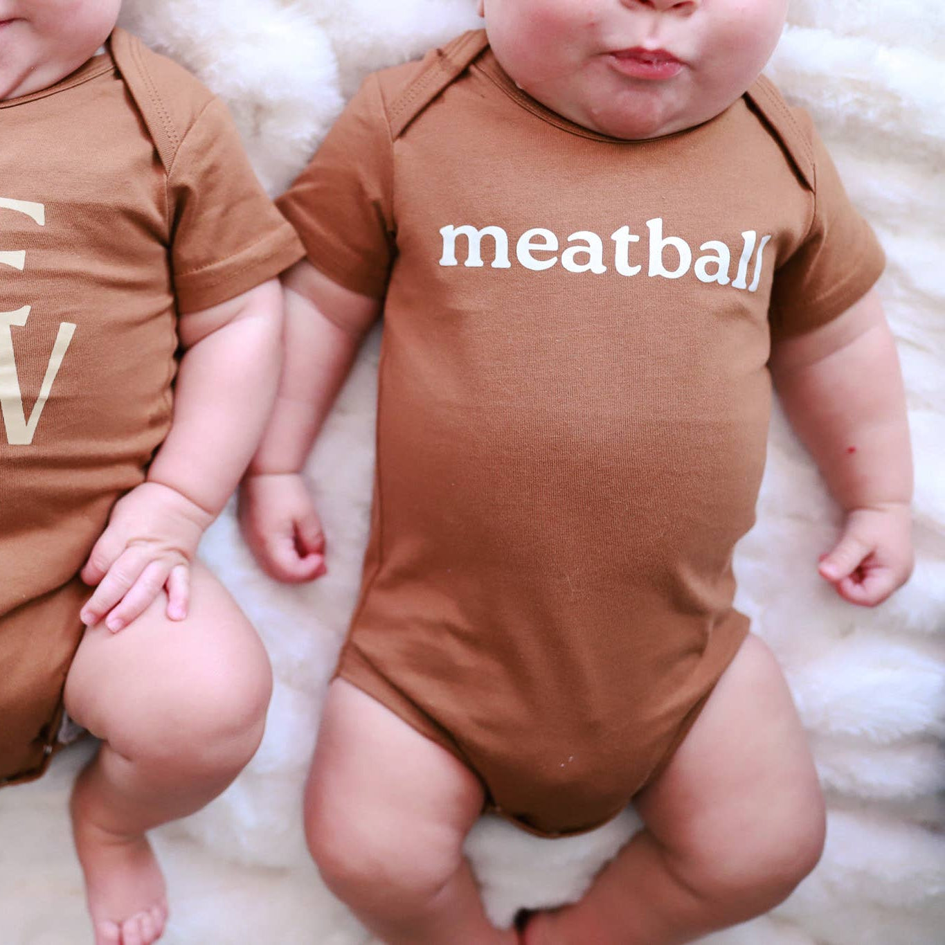 Two babies wearing brown onesies with text, sitting on a white surface.