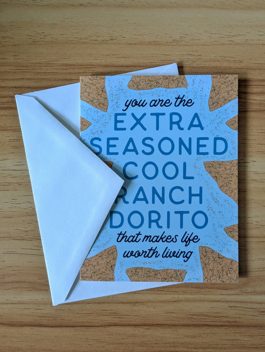 Card with blue text on a white envelope featuring a humorous message, "you are the extra seasoned cool ranch dorito that makes life worth living" on a wooden surface.