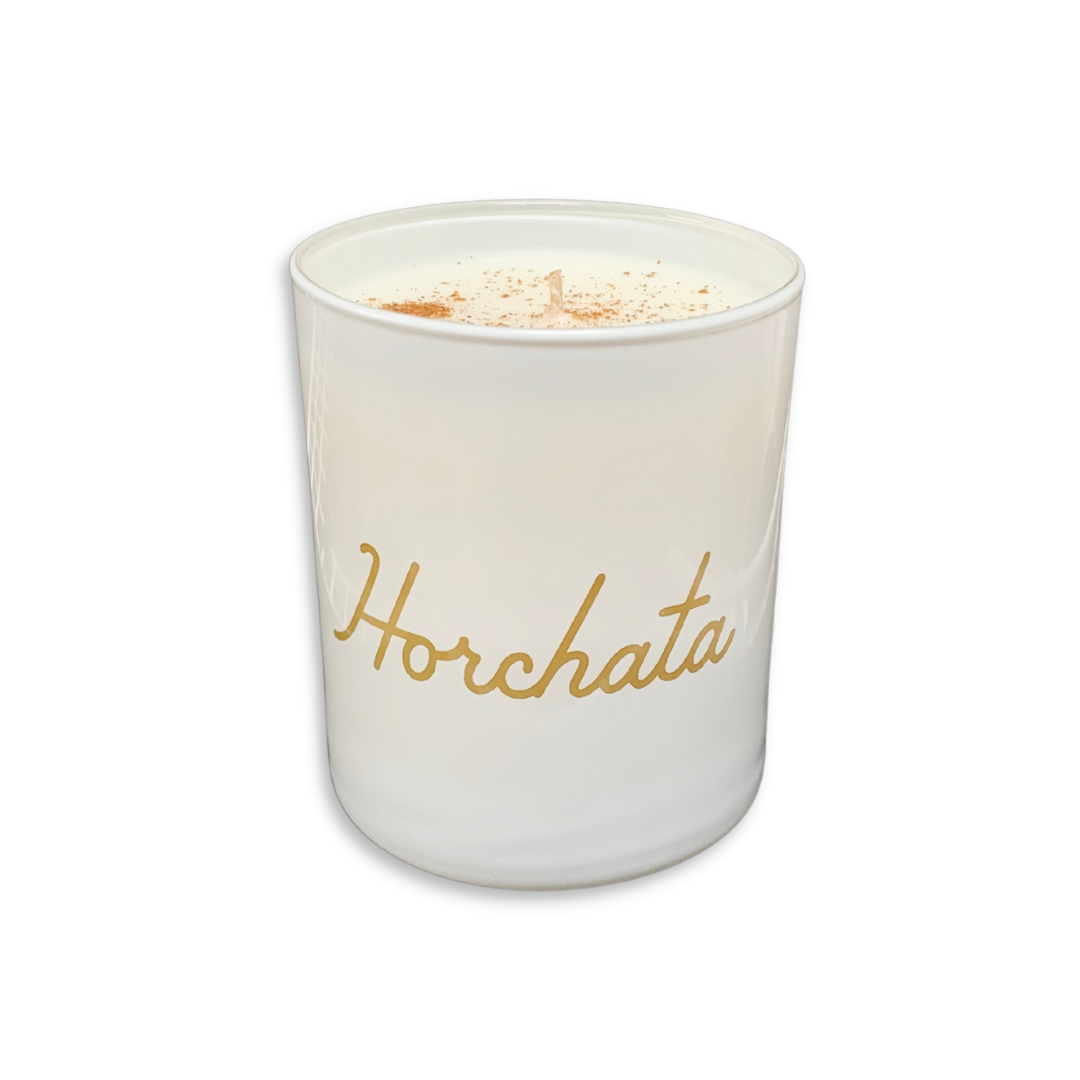 A white glass jar candle with the word 'Horchata' written in gold cursive script on the front, topped with white wax and a sprinkle of cinnamon, shown on a plain white background.