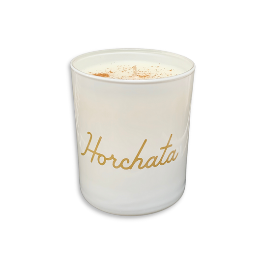 A white glass jar candle with the word 'Horchata' written in gold cursive script on the front, topped with white wax and a sprinkle of cinnamon, shown on a plain white background.