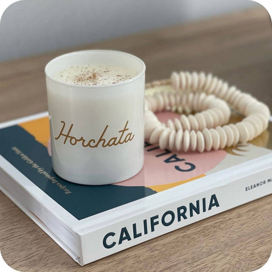 A white candle jar with the word 'Horchata' in gold script sits on top of a book, with a coiled cream cord and a wooden tabletop visible around it.