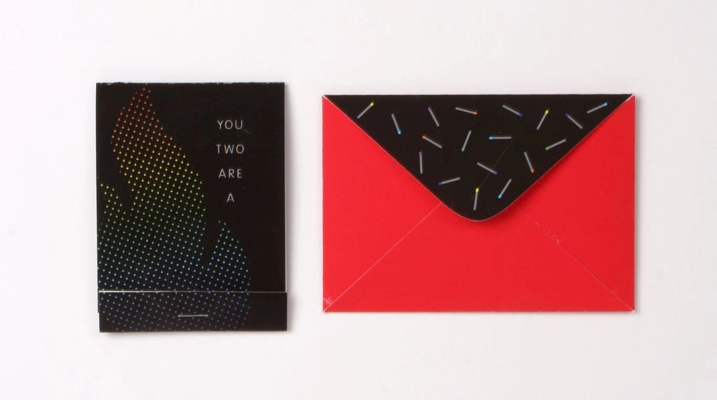 Photo showing front of matchbook greeting card - black with subtle flame design and text "You Two Are." Next to the card is a red envelope (shown from behind) with black flap and rainbow colored matches printed onto flap.