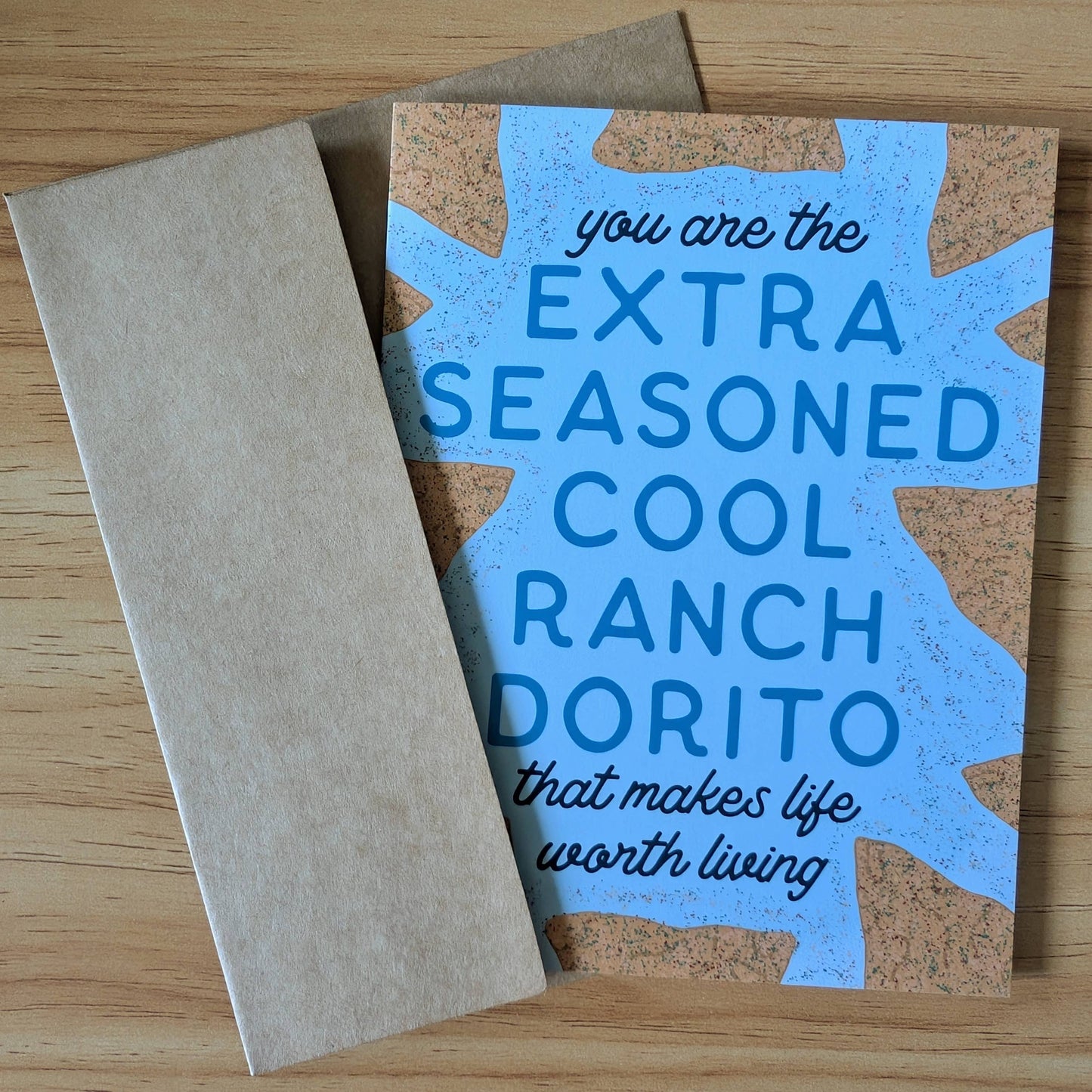 Card with blue text on a brown envelope featuring a humorous message, "you are the extra seasoned cool ranch dorito that makes life worth living" on a wooden surface.