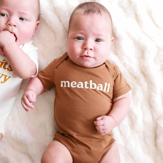 Baby wearing a brown onesie with 'meatball' printed on it, lying on a white fluffy blanket.