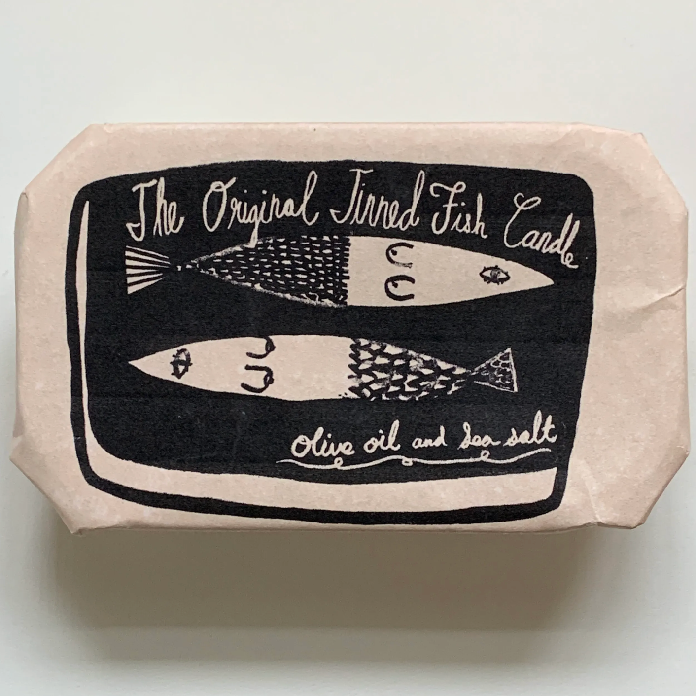 Tinned Fish Candle outer paper packaging -- text up top reads "The Original Tinned Fish Candle", two fishes in the center and underneath text reads "olive oil and sea salt" 