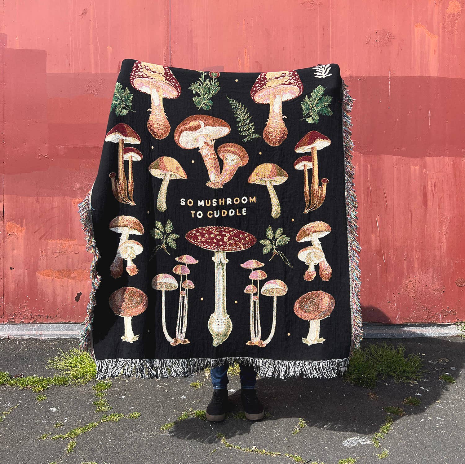 Mushroom blanket held up in front of red wall. Blanket has mushrooms and foliage on it along with text that reads "So Mushroom To Cuddle" 