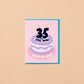 A square birthday card with a pink front and turquoise border, featuring a stylized purple cake with the text '35 Still Alive!' and 'Happy Birthday!' on a warm beige background.