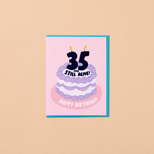 A square birthday card with a pink front and turquoise border, featuring a stylized purple cake with the text '35 Still Alive!' and 'Happy Birthday!' on a warm beige background.