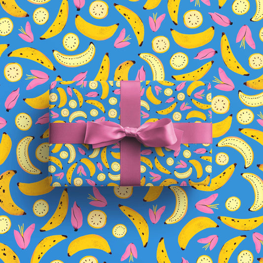 Gift wrap paper sheet with already wrapped gift on top. Blue gift wrap sheet with yellow bananas, both closed and sliced open, along with pink flowers on it 
