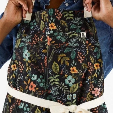 Details of apron that is mostly dark with lighter colored florals, herbs and foliage on it. Cream colored neck loop + tie 