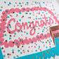 Congrats sheet cake card with sprinkles and confetti. The cake has a slice taken out of it to reveal chocolate cake with pink frosting. Close up photo to show the foil detail of the confetti.