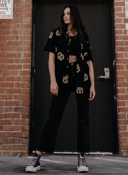Photo of woman with long hair wearing pretzel shirt unbuttoned over black crop top, with black jeans and sneakers.