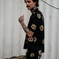 Photo of man with tattoos on his arm wearing button up pretzel shirt and black jeans.