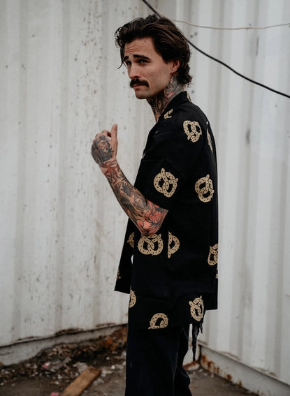 Photo of man with tattoos on his arm wearing button up pretzel shirt and black jeans.