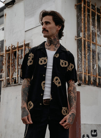 Photo of man with tattoos on his arms wearing pretzel shirt, unbuttoned over a white undershirt and black jeans.