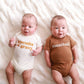 Two babies wearing onesies with text, lying on a white textured surface.
