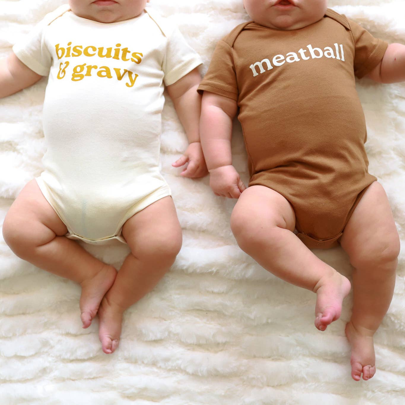 Two babies wearing onesies with text, lying on a white fluffy surface.