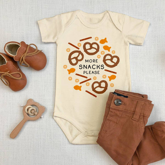 Flat lay of a baby onesie that reads "more snacks please" in the center of various snacks including cheerios, pretzels and goldfish, along with brown baby pants, shoes and a wooden toy