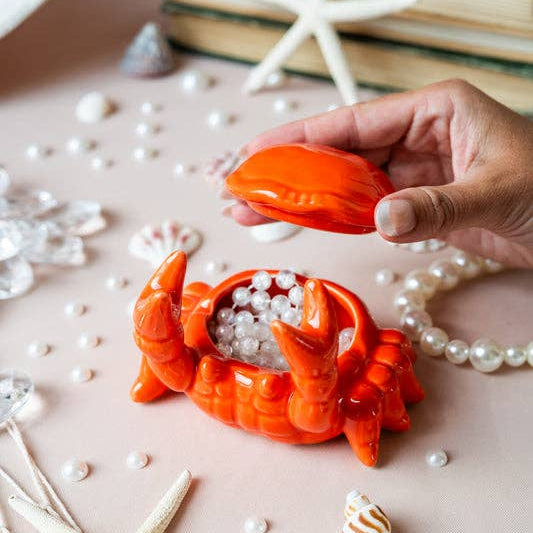 Hand holding a red crab-shaped object with pearls on a pink surface with starfish and books.