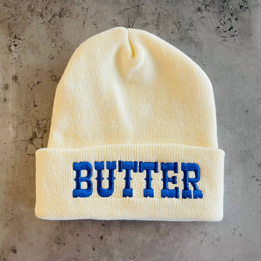 Ivory knit beanie with "BUTTER" in blue embroidery.