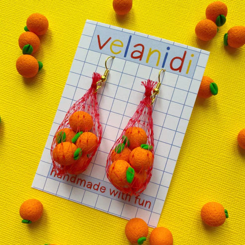 Oranges in mesh bag earrings on card backing that reads "velanidi handmade with fun" All on yellow background with scattered mini clay oranges on it 