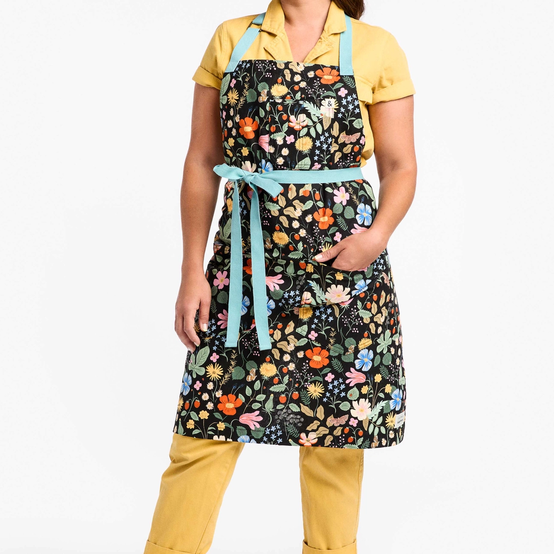 Model wearing apron with colorful design pattern of bright florals and foliage and light blue neck and waist straps 