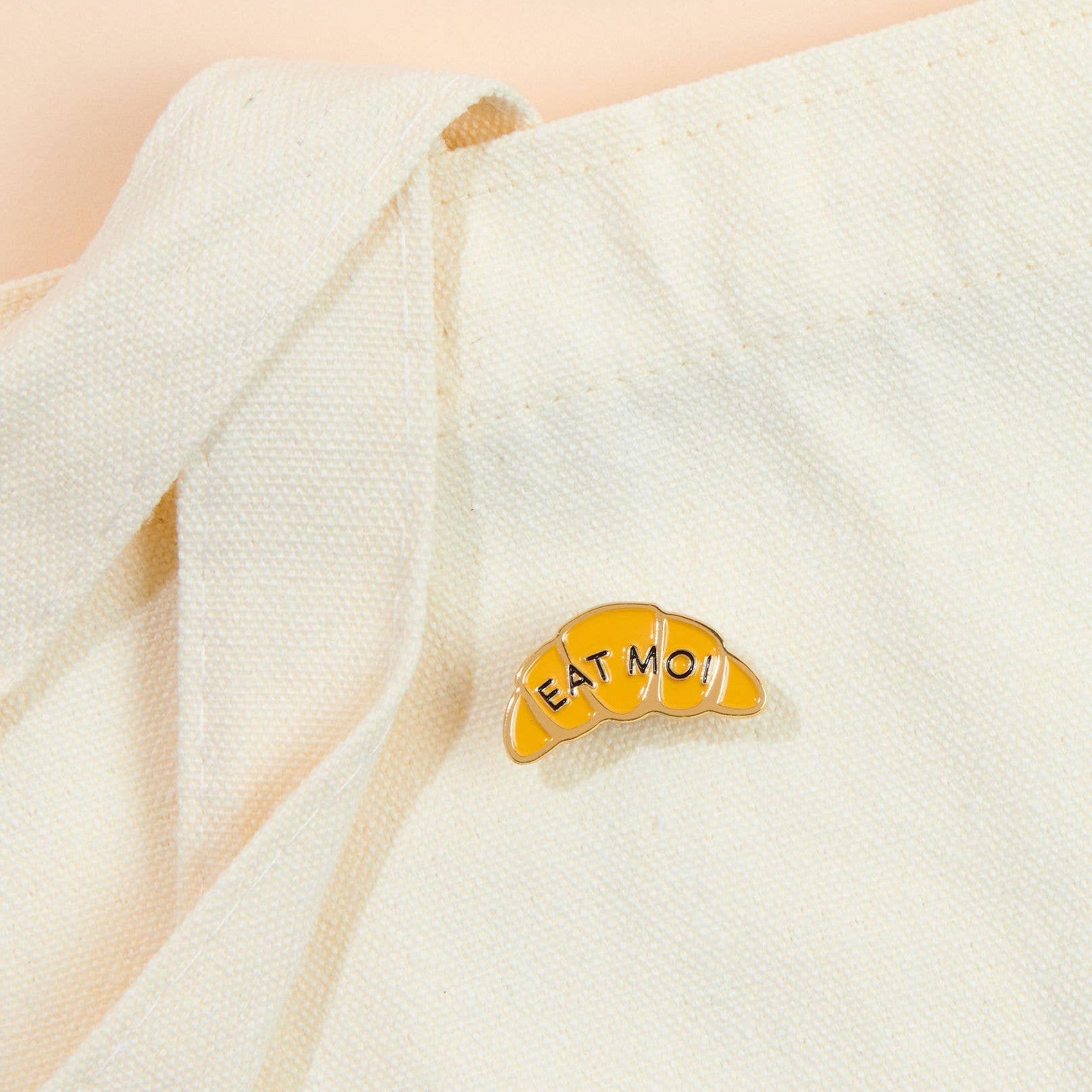 Enamel pin in the shape of a croissant that says "EAT MOI". Staged on a beige tote bag.