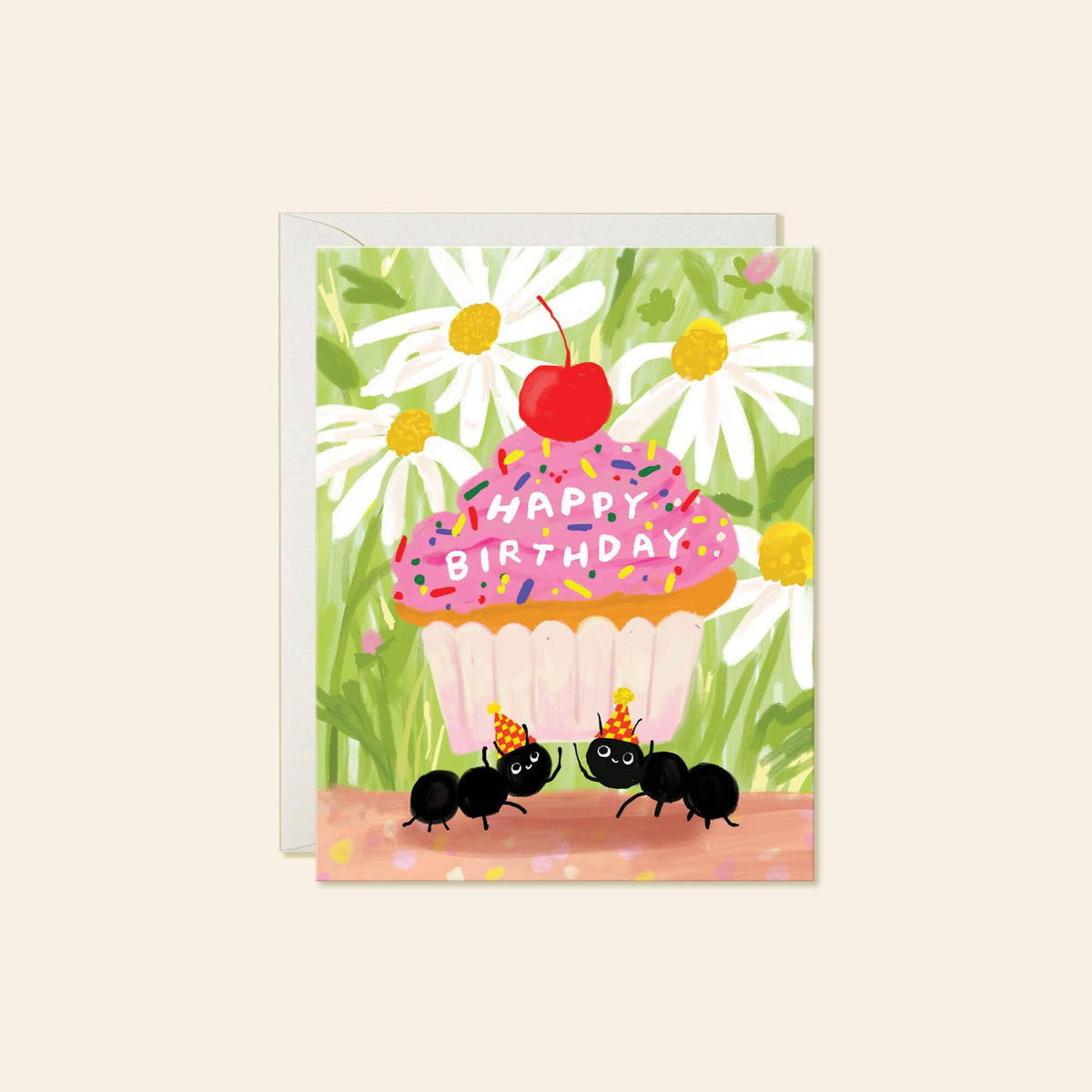 A square birthday greeting card with a white envelope behind it, featuring a colorful illustration of a cupcake with pink frosting, a red cherry on top, white daisies, green leaves, and two black ants wearing party hats. The card reads 'HAPPY BIRTHDAY' across the cupcake.