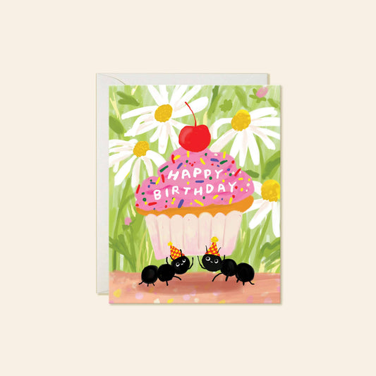 A square birthday greeting card with a white envelope behind it, featuring a colorful illustration of a cupcake with pink frosting, a red cherry on top, white daisies, green leaves, and two black ants wearing party hats. The card reads 'HAPPY BIRTHDAY' across the cupcake.