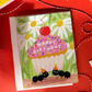 A square birthday greeting card with a white envelope behind it, featuring a colorful illustration of a cupcake with pink frosting, a red cherry on top, white daisies, green leaves, and two black ants wearing party hats. The card reads 'HAPPY BIRTHDAY' across the cupcake.