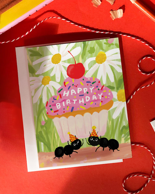 A square birthday greeting card with a white envelope behind it, featuring a colorful illustration of a cupcake with pink frosting, a red cherry on top, white daisies, green leaves, and two black ants wearing party hats. The card reads 'HAPPY BIRTHDAY' across the cupcake.