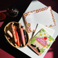 A square birthday greeting card with a white envelope behind it, featuring a colorful illustration of a cupcake with pink frosting, a red cherry on top, white daisies, green leaves, and two black ants wearing party hats. The card reads 'HAPPY BIRTHDAY' across the cupcake.