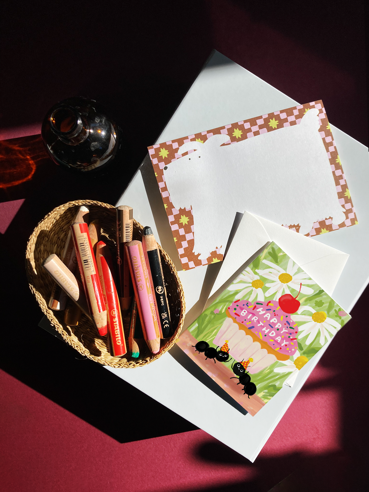 A square birthday greeting card with a white envelope behind it, featuring a colorful illustration of a cupcake with pink frosting, a red cherry on top, white daisies, green leaves, and two black ants wearing party hats. The card reads 'HAPPY BIRTHDAY' across the cupcake.