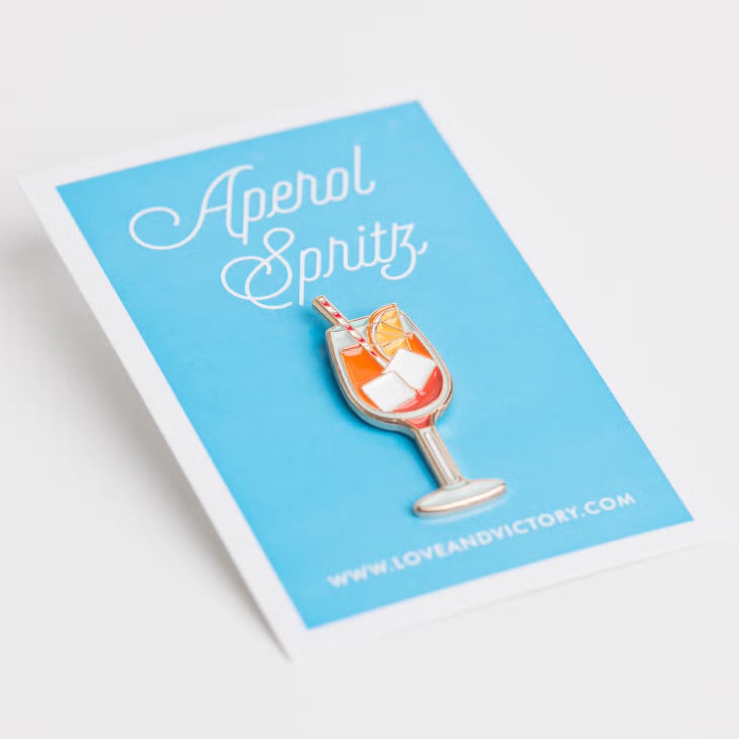 Aperol Spritz lapel pin on baby blue card backing.