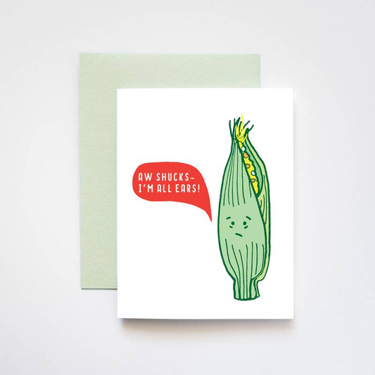 A white greeting card with a humorous illustration of a corn cob with a red speech bubble that reads 'AW SHUCKS - I'M ALL EARS!' in white text, shown with a pale green envelope on a light gray background.