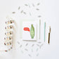A styled flat-lay on a white background featuring a greeting card with a cute cartoon corn illustration and the text 'AM SICKER, I BLOOM!'. Surrounding the card are silver paperclips, a roll of washi tape with colorful bird illustrations, a white pen, a mint green striped pen, and a gold pen.