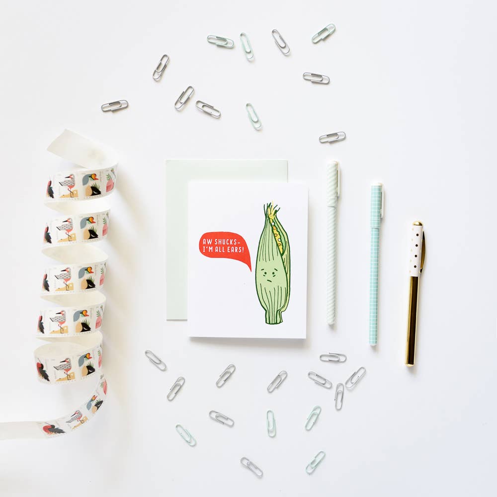 A styled flat-lay on a white background featuring a greeting card with a cute cartoon corn illustration and the text 'AM SICKER, I BLOOM!'. Surrounding the card are silver paperclips, a roll of washi tape with colorful bird illustrations, a white pen, a mint green striped pen, and a gold pen.