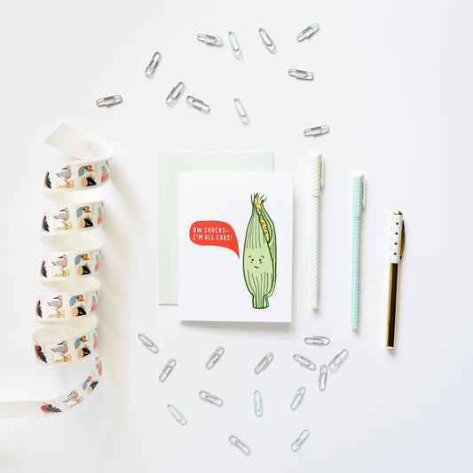 A styled flat-lay on a white background featuring a greeting card with a cute cartoon corn illustration and the text 'AM SICKER, I BLOOM!'. Surrounding the card are silver paperclips, a roll of washi tape with colorful bird illustrations, a white pen, a mint green striped pen, and a gold pen.