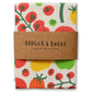 A folded cotton tea towel with a colorful tomato print, wrapped in a brown paper band that reads 'BADGER & BURKE 100% COTTON TEA TOWEL'.