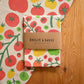 A folded cotton tea towel with a colorful tomato print is wrapped in a brown paper band labeled 'BADGER & BURKE'. The towel is displayed on a wooden surface next to a larger matching flat lay of the same towel.