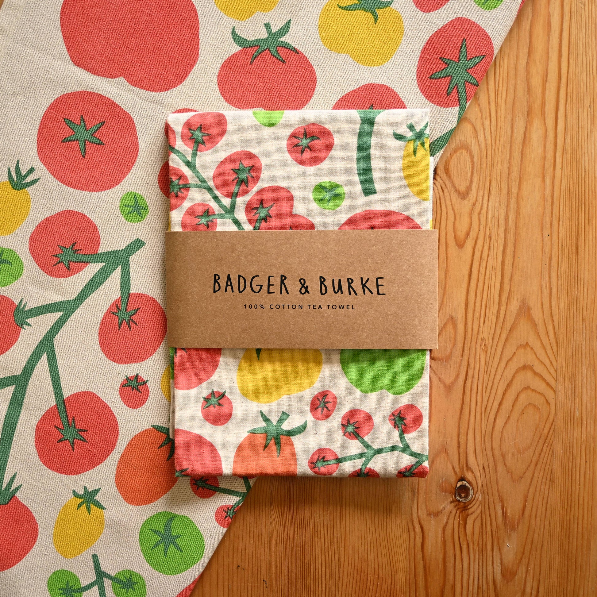 A folded cotton tea towel with a colorful tomato print is wrapped in a brown paper band labeled 'BADGER & BURKE'. The towel is displayed on a wooden surface next to a larger matching flat lay of the same towel.