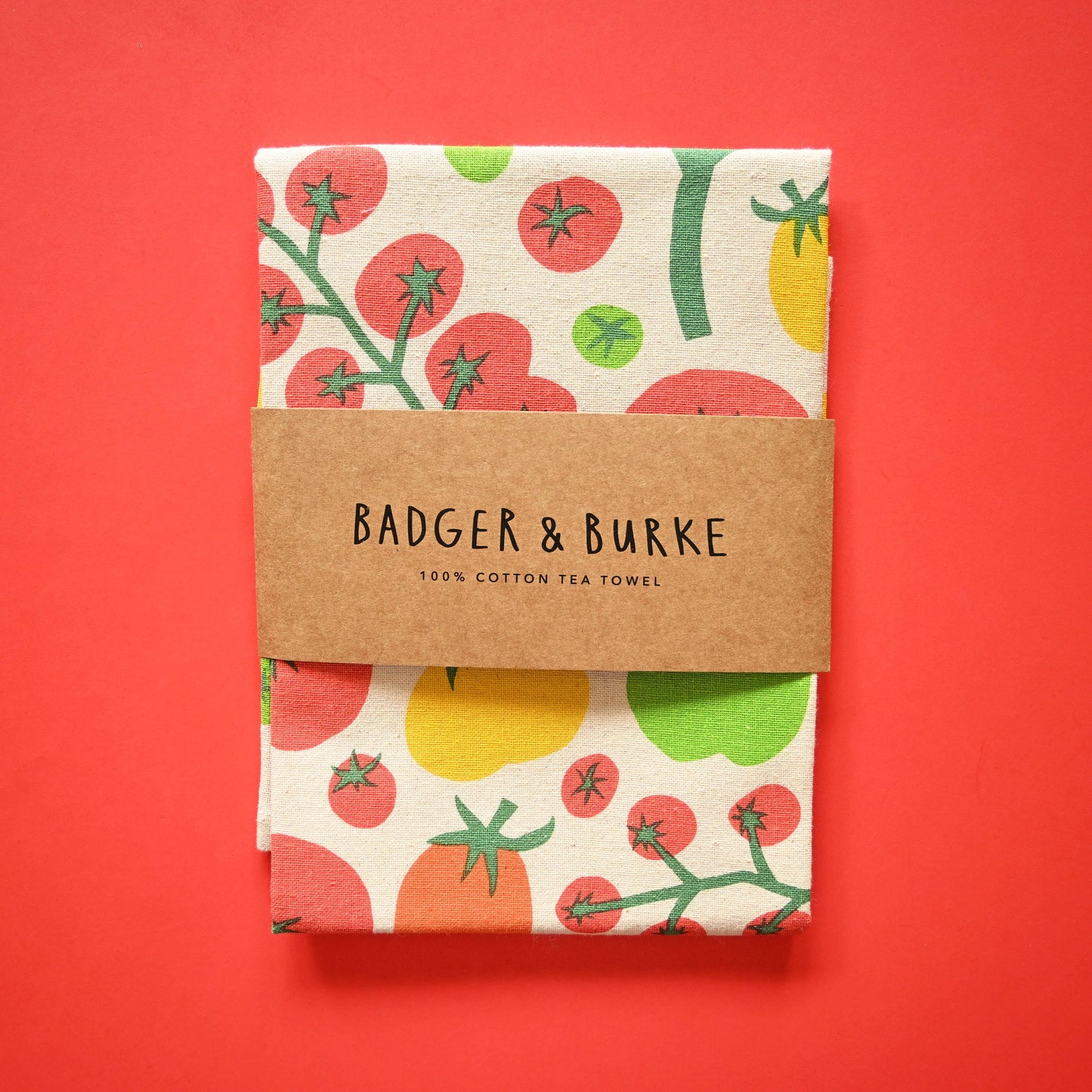A folded cotton tea towel with a colorful tomato and leaf pattern, wrapped in a brown paper band that reads 'BADGER & BURKE 100% COTTON TEA TOWEL', set against a solid red background.