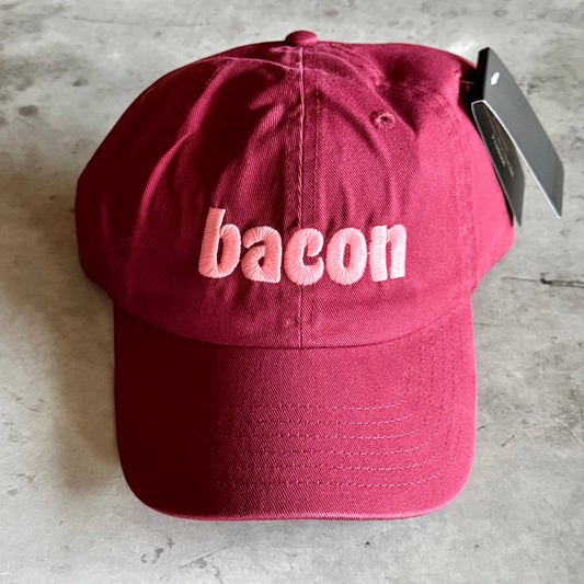 A dark red baseball cap with the word 'bacon' embroidered in light pink across the front, resting on a gray concrete surface with a black retail tag attached.