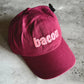 A burgundy cotton baseball cap with the word 'bacon' embroidered in light pink across the front, resting on a gray concrete surface.