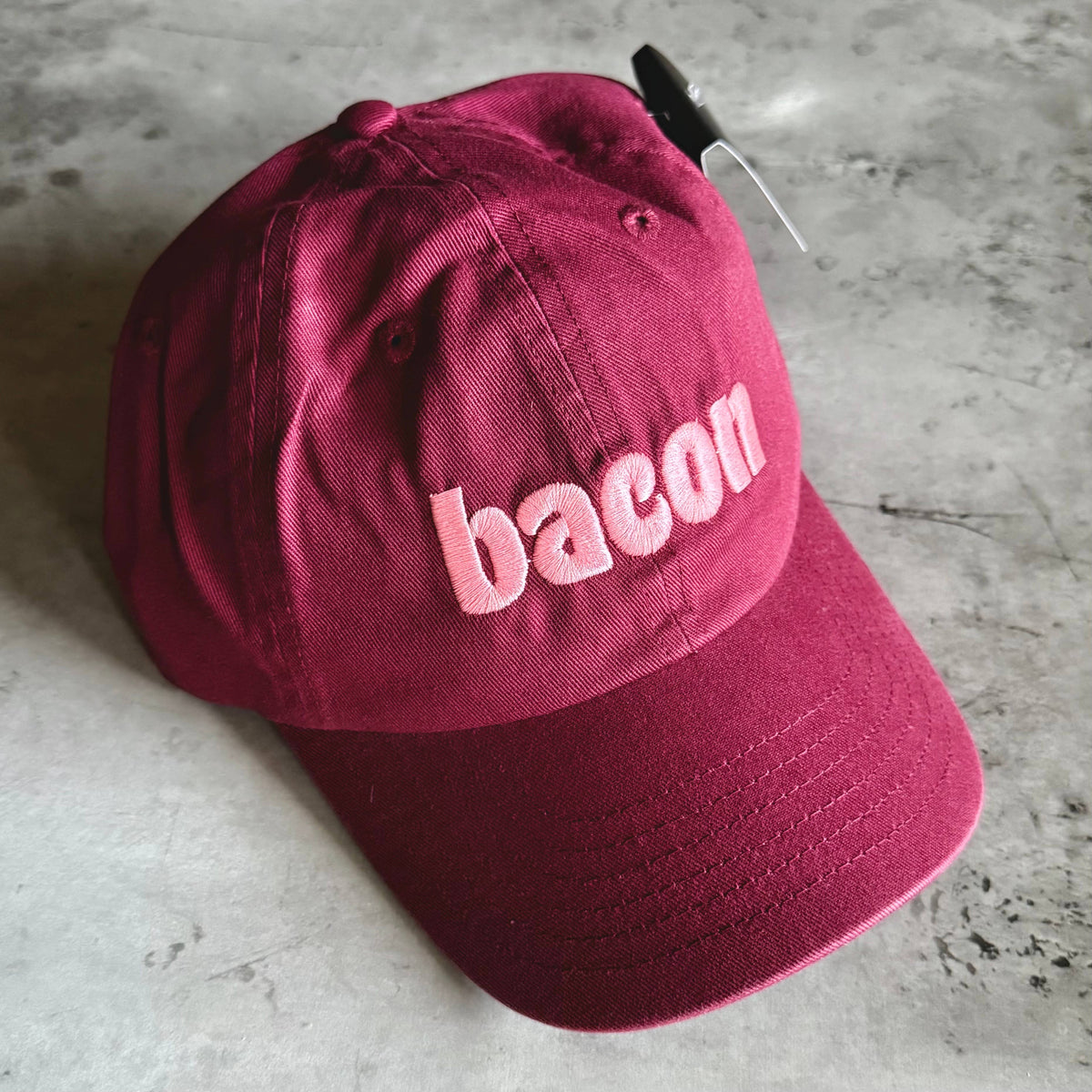 A burgundy cotton baseball cap with the word 'bacon' embroidered in light pink across the front, resting on a gray concrete surface.